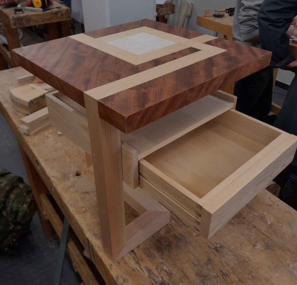 Photo d'une petite table basse en bois au design pleins de carré. Le plateau et le piètement forme le volume d'un cube qui n'est pas plein. Le plateau est marqueté avec des bandes de bois clairs qui viennent prolonger les angles formés par les pieds. Sous le plateau il y a un tiroir suspendu