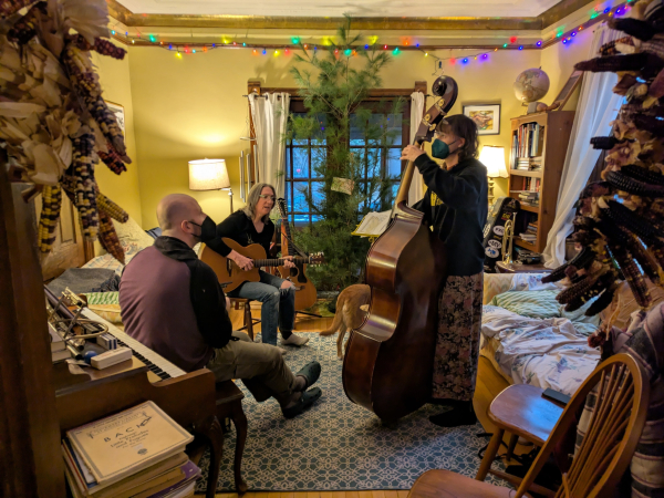 Small parlor, shot from just outside the double door frame. Me, on acoustic guitar, center-left, facing the camera, near the back, an unadorned solstice tree behind me, and French doors to the porch behind all; middle aged white woman playing upright bass standing by an overstuffed couch, foreground right; middle aged white guy seated by the piano, foreground left, playing mandolin. Bass and mando players are 3/4 profile away from the camera.

Colored fairy lights encircle the room, just below the high ceiling.

Dried flour corn hangs from the door frame, just in front of the camera.

Edit: the hind end of a cute blonde doggo is just visible behind the bass.