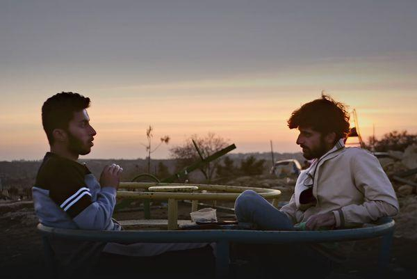 A still from the film No Other Land. Two men of Middle Eastern appearance sit and talk on a roundabout in a children's playground at sunset, with open country stretching out to the horizon behind them.