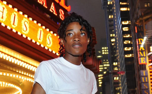 Bust shot of Jordan Neely dressed as Michael Jackson, with dapper curls and a white t-shirt, standing in front of a theater marquee on a busy downtown street at night. He’s a thin black man and his face looks of contentedness and comfort.