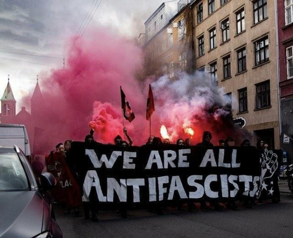 Foto eines schwarzen Banners, auf einer Demo, auf welchem „we are all antifascists“ steht. Hinter dem Banner zu sehen einige vermummte Menschen mit zwei Fahnen und roter Pyro