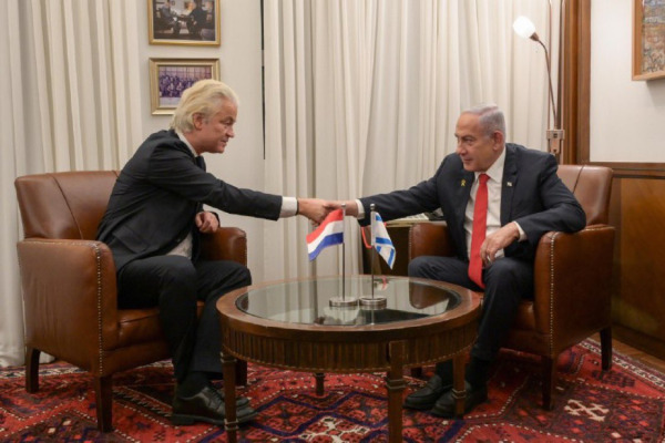 Wilders schudt de hand van Netanyahu terwijl ze ieder in een fauteuil zitten. Op de ronde salontafel voor hen staan twee vlaggenstandaardjes: één met de Israelische vlag en één met de Nederlandse vlag.