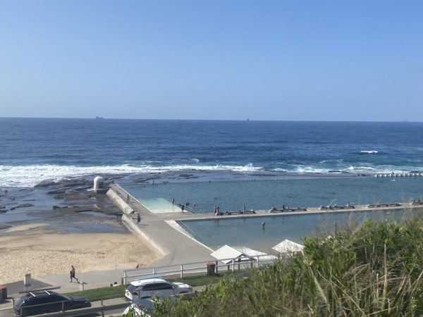 Looking down at the ocean baths at out to sea from the hill above on a sunny day. Lots of blue water and sky. Both pools visible, with the rounded white pump house in the far left corner and the white concrete lap lane markers on the far right of the main pool. Close to low tide so rocks exposed behind and to the side of the pools 