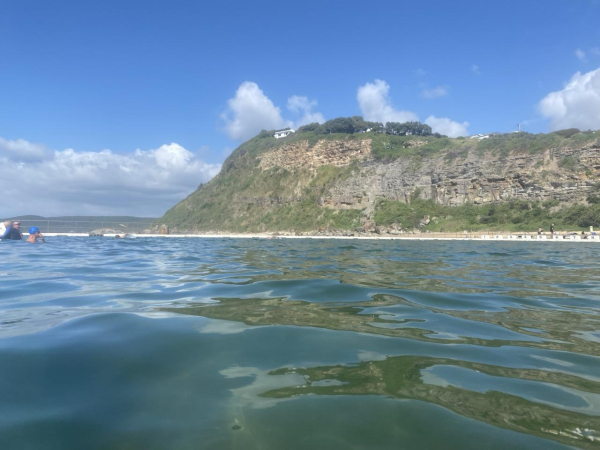 Taken while swimming so the blue green water is first, reflecting pieces of overgrown rocky bluff above. Puffy white clouds cling to the coastline but there's plenty of bluesky above. A couple of lap swimmers are resting on the far left just close enough to make out that one is in a black rashie and the other has a blue swim cap showing. 