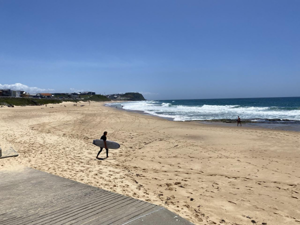 Surfer with a white board walking across the sand towards the waves. Light golden sand, vivid blue-green ocean and light blue sky.