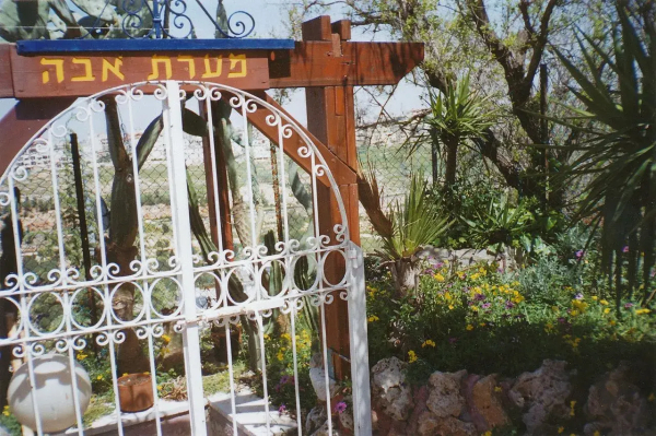 The image features a decorative gate or entrance with a sign above it that appears to be in Hebrew. The area around the gate includes greenery, including plants and flowers, suggesting a garden or rural setting. The gate has a white metal design, and there are various plants, possibly cacti or succulents, visible in the background.

Image Credits: Wikimedia / משה גלנץ / CC BY-SA 3.0