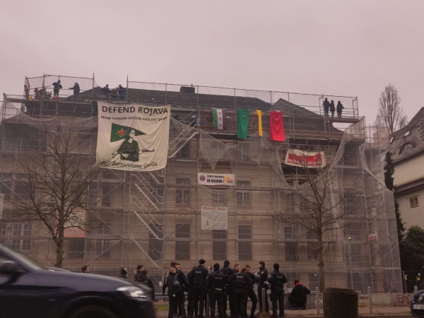 Eine neoklassizistische Villa mit vier Stockwerken ist von einem Baugerüst umgeben. 
An diesem hängen verschiedene Banner, eine aktuelle Flagge Syriens und Rojavas sowie farbige Tücher (grün, gelb, rot). 
Oben auf dem Baugerüst befinden sich Menschen. 
Im Vordergrund steht eine Gruppe Polizist*innen. 