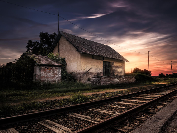 The photograph captures a rustic scene at sunset, near a small railway station in a rural part of Hungary. In the foreground, aged train tracks stretch towards a weathered, old building. The building appears abandoned, showing signs of decay with peeling paint and a worn roof. Behind it, a smaller, brick structure seems to be attached or perhaps a separate outbuilding. The background is a mix of vegetation, trees silhouetted against the vibrant sunset hues, and a clear, partly cloudy sky. The light suggests it is late in the day, casting long shadows and creating a dramatic contrast between light and dark tones.