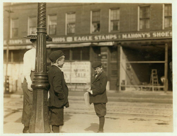 The image is a black and white photograph depicting two young boys dressed in early 20th-century attire, standing on a street corner. One boy appears to be holding some papers or tickets while looking towards the camera with a serious expression; he wears a dark jacket, shorts, socks, shoes, and a flat cap. The other boy is turned away from the camera, wearing similar clothing but without any visible accessories.

In the background, there are various signs for local businesses including "Eagle Stamps" and "Mahony's Shoes," indicating that they're on Grand Avenue in downtown St. Louis, Missouri during May 9th, 1910 as indicated by a caption below the image. A street lamp post is visible near them with text inscribed along its side.

The overall scene suggests a snapshot of daily life and work for young boys at this time period within an urban environment in America's heartland city.