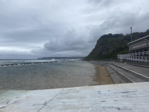 Looking across the shallower pool at Merewether Ocean Baths and southerly towards the overgrown rocky bluff and curve of coastline beyond. It's a cool day (23C max) after heatwave conditions yesterday (38C max). Grey-tinged white clouds fill the sky. The shallow pool looks grey and subdued. 