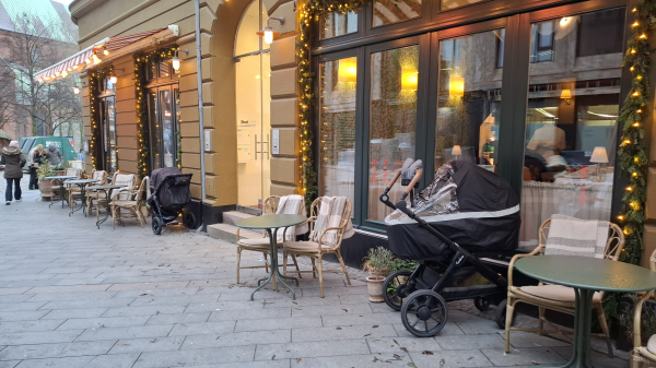 One of the main streets in the very centre of Aarhus, Denmark. Early afternoon. We can see a glimpse of the redbrick, green-roofed cathedral ahead and a few pedestrians. To our right, café windows with Christmas decorations and the blurred figures of people inside. Outside, pavement tables and chairs with blankets for keeping you warm, though nobody is sitting outside right now. In front of each of two separate café sections, outside the window on the street, a loan black pram with unseen babies sleeping quietly and happily and peacefully inside. This is not uncommon in Scandinavia. Parents can see them still and if a passerby does hear a child wake up and start to cry before the parent does, they’ll just stick their head into the café and pass on this breaking news.