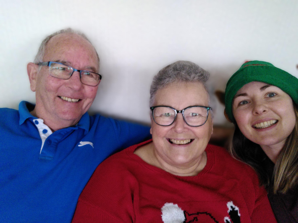 Three people all related. Mother and father and adult daughter. All smiling, mother is wearing red Christmas jumper and chemo curls. Daughter is wearing elf ears.