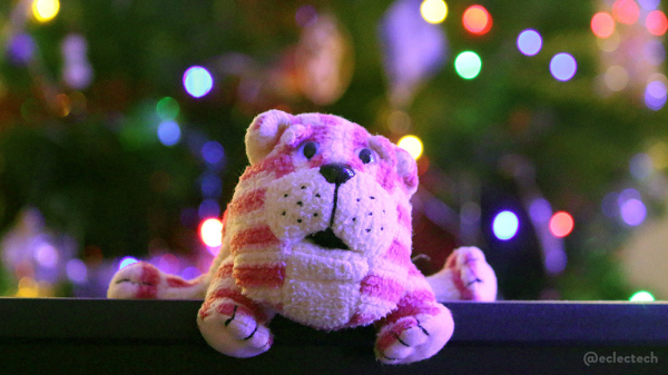 A photo of a small Bagpuss beanbag toy sitting on the top of a black monitor. Behind in soft focus is a Christmas tree shining with lights and baubles. Bagpuss is a pink and white stripey cat from an old, very good, kids TV show in the UK.