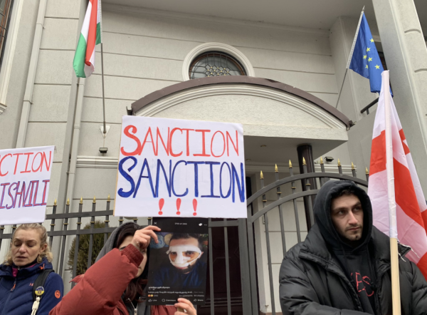 Protestors outside the Hungarian Embassy in Tbilisi, Georgia. They have Georgia flags and a sign saying 'Sanction Sanction'