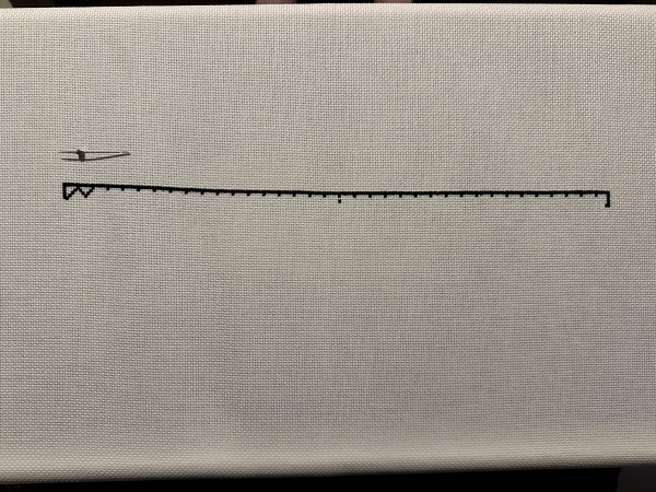 A long line of black cross stitch. There a needle for scale. It’s an out 13 inches long. A zigzag pattern under the line is beginning to form. 