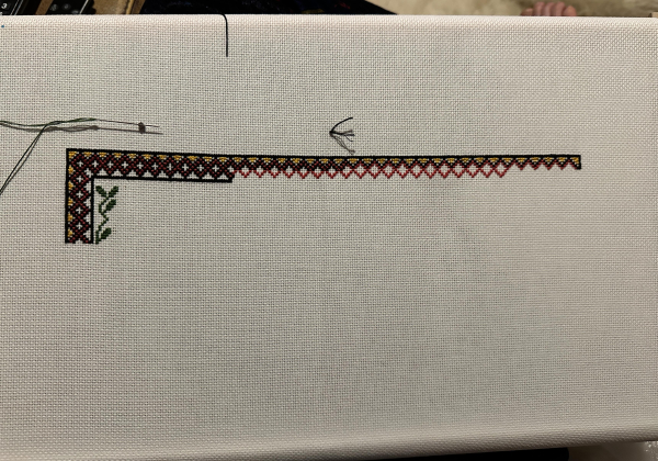 A partially completed cross stitch border in yellow, black and red. In the left hand top corner are a few simplified cross stitch leaves. 