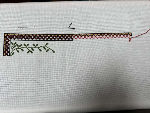 A cross stitch sampler in the middle of being made. A partially stitched border in black, red and yellow hors across the top. Small leaves stretch across from the left hand side. 