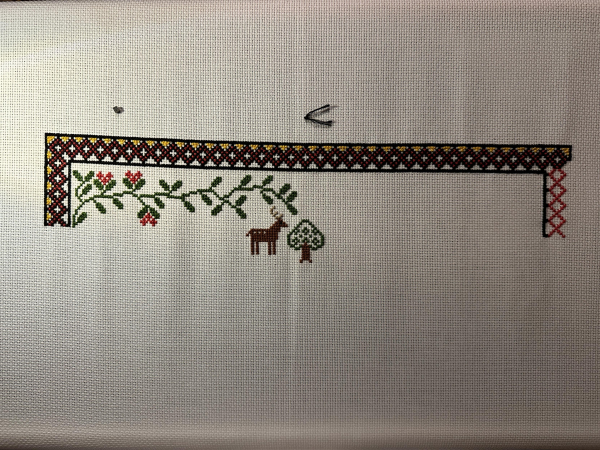 Cross stitch sampler work in progress. Border on the left and top is done, but the right hand side is incomplete. Some red flowers are on the foliage under the border. A stag and a small tree are in the middle. 