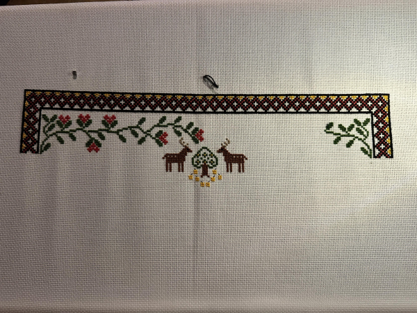 Partially completed cross stitch sampler. Border on the left, top and right. Foliage on the left and right. Two stags and a tree in the middle.