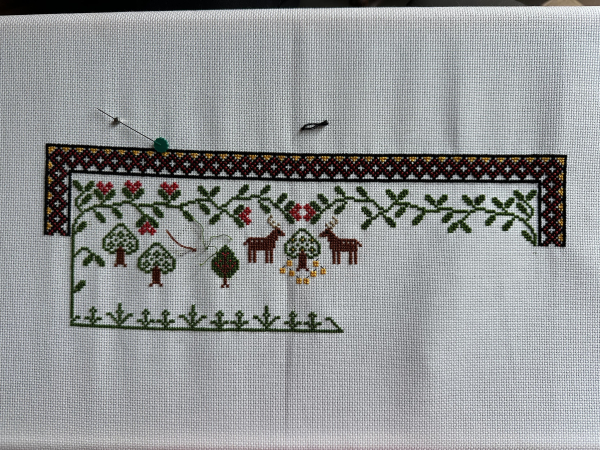 Cross stitch sampler in the process of being made decorative border, foliage, trees and stags are visible. And at the bottom there are some stalks which will have flowers on them. 