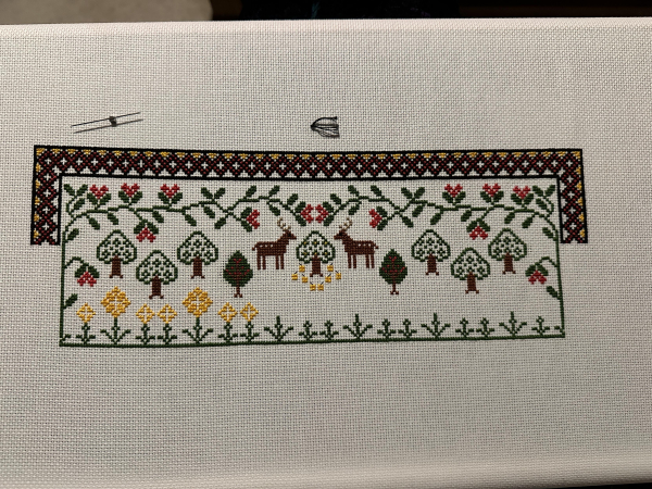 Cross stitch sample work in progress. Stylised leaves, tree and stags are on show with a decorative border on the top and the sides. At the bottom are some stalks. Some of them have flowers on.