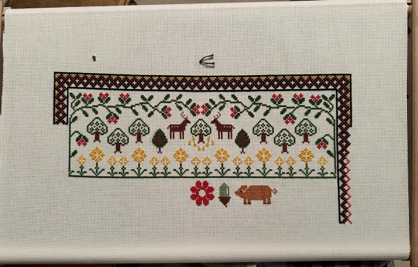 A work in progress cross stitch sampler. Flowers, leaves, trees and flowers on the top. A large red flower, an acorn and an orange pig underneath it. A partially completed decorative border goes around the top and down the right hand side. 