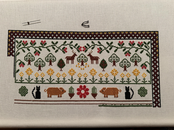 Cross stitch sampler. Moving down it: Stylised forest scene at the top. Then some yellow flowers. A row of four leaf clover, cats, pigs and acorn. Part currently being worked on is a wooden fence and grass 