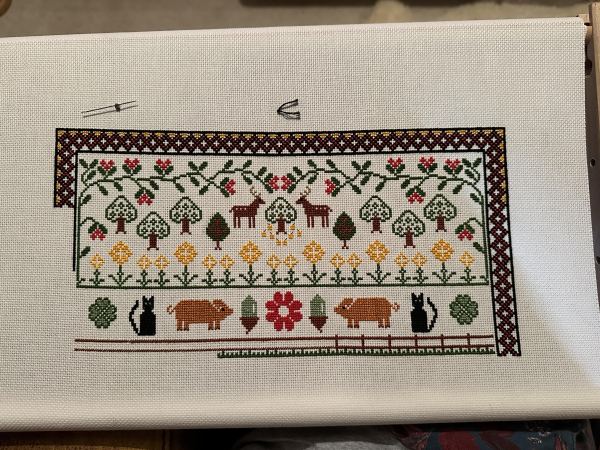 Partially completed cross stitch sampler. Stylised forest and deer at the top, pigs, cats, acorns and clover at the bottom. At the very bottom is a fence which hasn’t been completed yet.