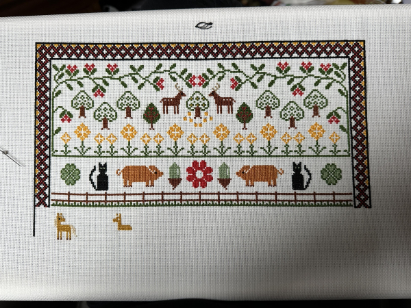 Work in progress cross stitch sampler. There are deer and a forest at the top. Then there are cats, pigs and acorns. Under that there is a palomino pony and another partially completed pony. Everything is stylised and the scale is odd. There’s also a decorative border on the left, top and right. 