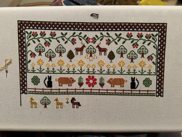 Partially completed cross stitch sampler. There are lots of animals, trees and flowers which are all stylised rather than realistic. The part currently being worked on has two Haflinger ponies and a bay. 