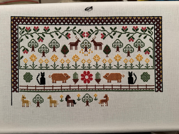 I work in progress Crosstitch sampler. At the top there are leaves and trees and flowers and deer. below that there is four leaf clover, cats, Pigs, acorns and a big flower. then there’s a fence. Below the fence are four ponies two Palominos then a bay and then a chestnut. There’s space on the right for two more ponies.