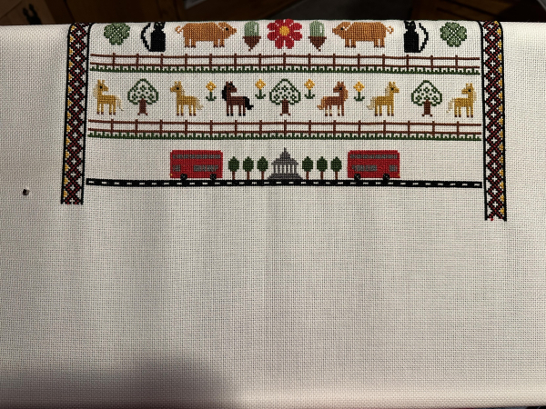 Cross stitch sampler work in progress. it features at the bottom two London double-decker buses which are on a road. they’re both facing away from each other but identical so nobody could ever get on the buses because there are no doors also in this section as university building with some trees.