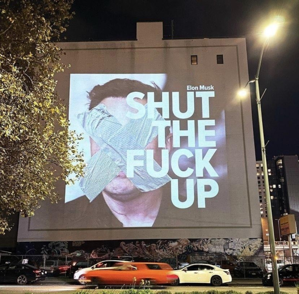 A projection on a building shows a large image of Elon Musk's face partially covered by grey duct tape. Overlaid on the image are the words "SHUT THE FUCK UP" in large, white, sans-serif font. The building is graffitied at the bottom. Several cars are parked in front of the building. A street light is visible to the right.

Provided by @altbot, generated using Gemini