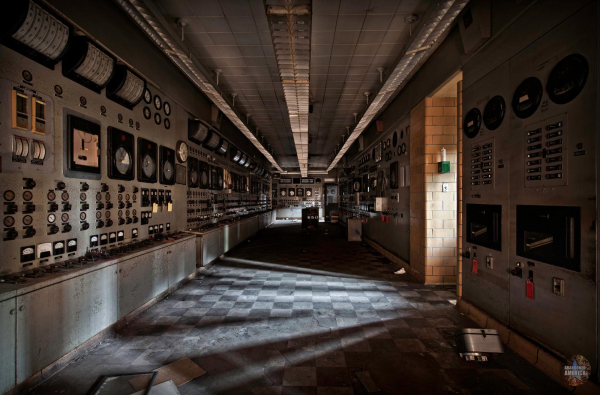 A long, dark room with a checkerboard pattern on the floor and rows of gauges, dials, and meters along the walls. A door on the right lets in a shaft of light