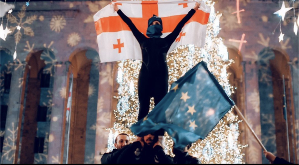 Khorumi dancers outside parliament in Tbilisi Georgia. A woman stands on the shoulders of a man. She is waving a Georgian flag and is silhouetted and illuminated by the lights of the Christmas tree and decorative lights. To the right an EU flag is waved.