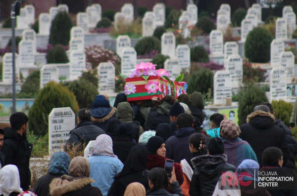 A crowd carrying the sarcophagus of Arab YPJ fighter Aruba Ali al-Kuno in the background are gravestones from the cemetery.