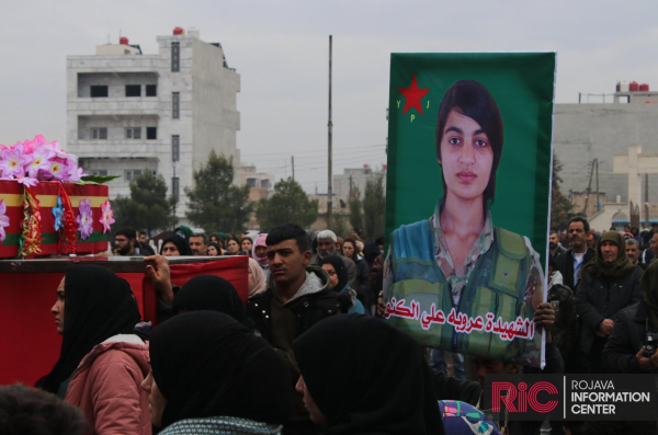 A crowd surrounds the sarcophagus of the Arab YPJ fighter next to a large photo of her face Aruba Ali al-Kuno in a YPJ uniform.