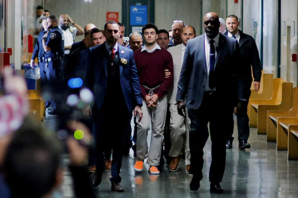 Mangione is led through the court house. He's dressed in a collard shirt and sweater. His hands and feet a shackled. He's surrounded by four police officers in formal wear and several uniformed police look on. 