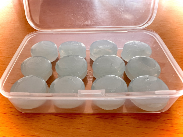 A clear plastic container filled with twelve round, gelatin-like earplugs shaped like lozenges. They have a translucent blue tint and are placed neatly inside the container.