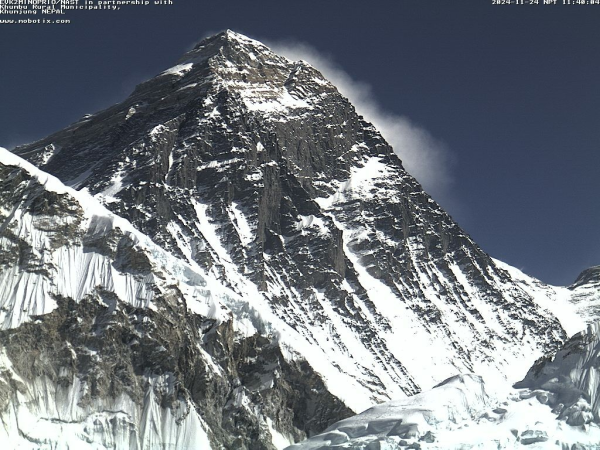 A photo captured by a webcam pointed at Mount Everest. Its largest peak is in the center of the image, rocky, sparsely covered by snow. Two shorter peaks are partially visible on either side.

Clear blue sky is visible behind the mountain.