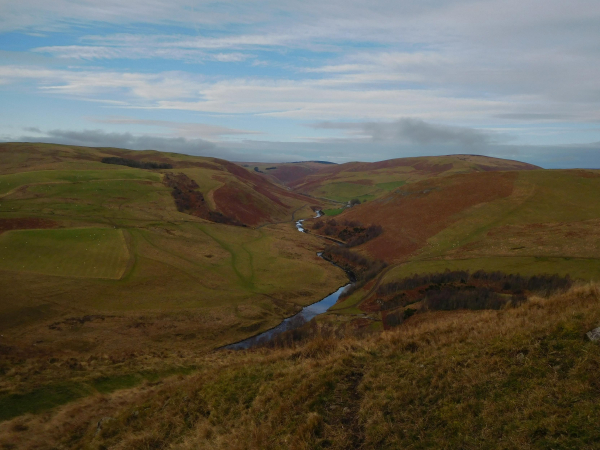 Colour photograph taken from a high advantage point looking down a long valley with gently sloping sides a river runs along the valley bottom. The sky is blue with white wispy clouds.