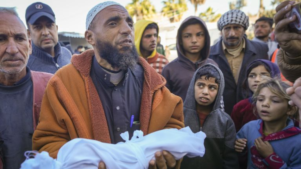 Imam Islam Abu Suaied met de overleden baby Jomaa al-Batran, in een wit doek gewikkeld, in zijn armen. Om hem heen staan Gazanen, die naar hem en de overleden baby in zijn armen kijken.