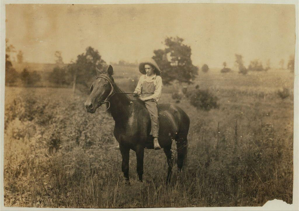 The image depicts a sepia-toned photograph of an individual riding on the back of a horse in what appears to be rural countryside. The person is wearing overalls and has their feet placed against the saddle, suggesting they are well adapted for outdoor activities or farm work. They wear a wide-brimmed hat that shields them from direct sunlight, indicating it may have been taken during daylight hours.

The horse looks healthy with its mane intact, hinting at proper care. The scene is set in an open field with tall grass and wildflowers swaying gently in the wind. In the background, several trees are scattered across rolling hills under a hazy sky that adds depth to the composition.

There's no modern technology or urban infrastructure visible; it suggests a time when horses were integral for transportation and labor on farms. The overall atmosphere evokes themes of simplicity, connection with nature, and perhaps an era where self-sufficiency was key in rural life.