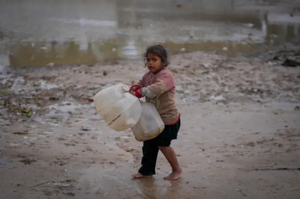 A young girl walks barefoot, carrying empty jerrycans to collect water, after overnight rainfall at the refugee tent camp for displaced Palestinians in Deir el-Balah [Abdel Kareem Hana/AP Photo]