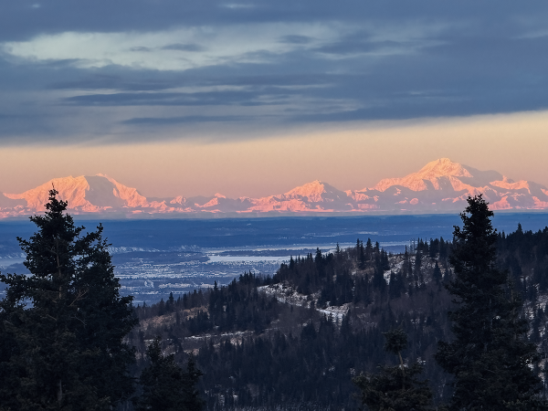 The Alaska Range glows golden and fills the horizon.  A broken layer of clouds above and blue tinged valley floor just behind a ridge in the foreground.