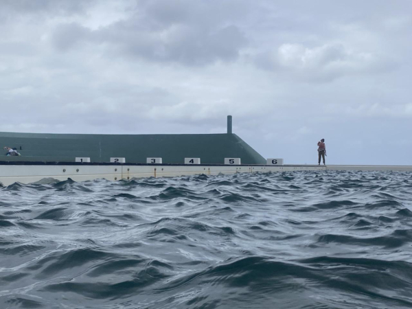 Taken while swimming on a cloudy day. There is choppy grey-green water in the corner of the main pool looking across to the lap pool and turquoise concrete wall bleachers. Six white concrete lap markers numbered in black are just visible above the wall of the pool. A person is standing to the right of the slanting end of the bleacher wall, holding a child. The clouds are white and grey.