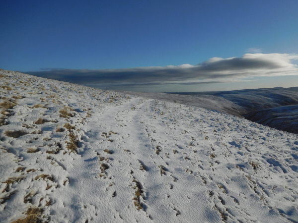 Colour photograph of a snowy slope running down from the left to the right of the frame. A faint track is just visible running away from the viewer. There are hills in the distance, in shadow. The sky is a deep clear blue at the top of the frame, but there is a line of grey cloud on the horizon.