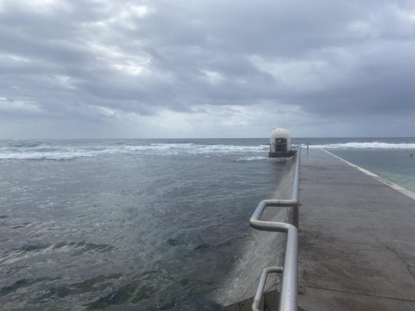 Looking down the stainless steel railing towards the rounded white pump house building and out to sea. On the right of the railing, there is the raised concrete concourse and the green-blue water of the main ocean pool. On the left, the concrete wall slants down to where the incoming tide has totally covered the natural rock platform. Further out, there's a line of white where the waves break on the edge of the hidden rocks. The sky is full of mostly grey clouds.