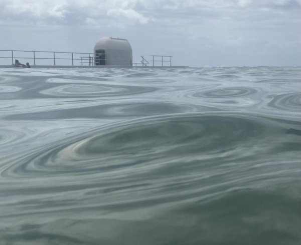 The surface of the undulating green water has formed concentric circular shapes. In the background, the rounded white pump-house and cloudy sky.