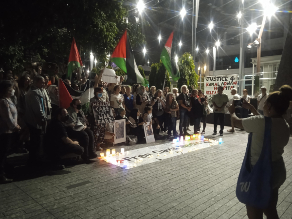 A group of health professionals stands in front of the "free Gaza medics" banner holding Palestinian flags and other signs with photos of Gaza medics